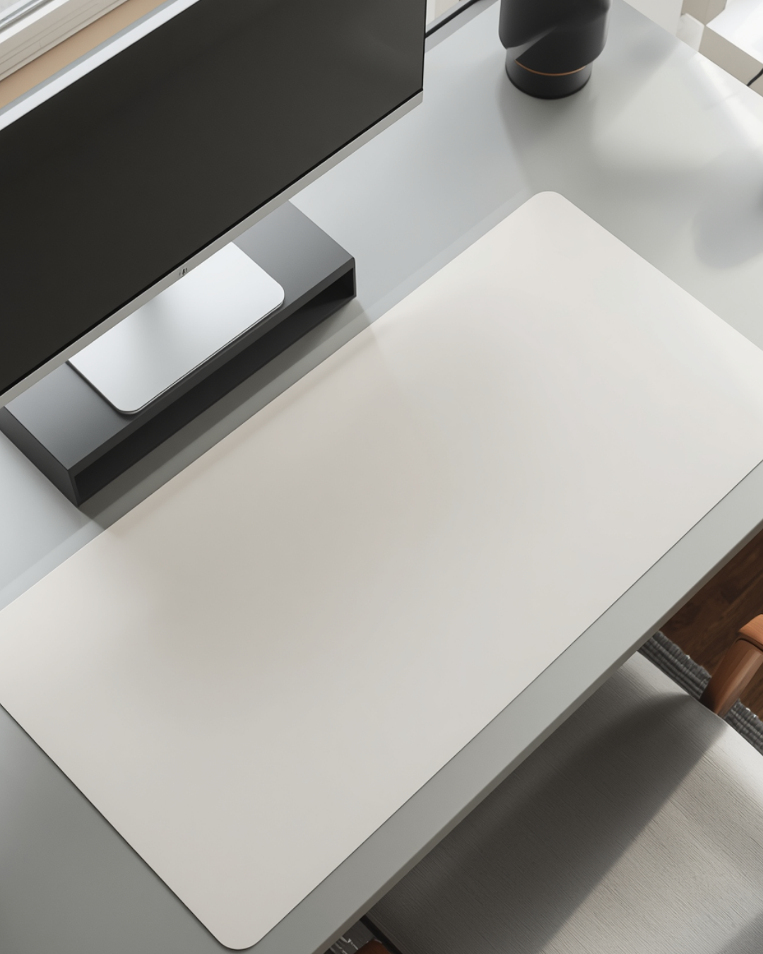 Top-down view of a tidy desk with a light desk mat and a monitor on a black riser, lit by window light